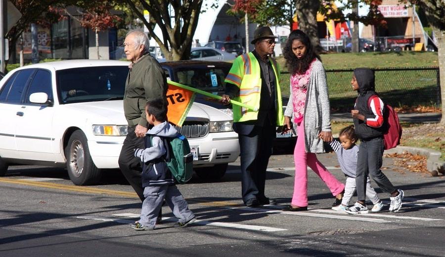 A group of students and families walk across the street as a crossing guard holds a stop sign to halt traffic